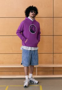 Sweatshirt roxa com capuz e logótipo, usada sobre uma camiseta riscada. Combinada com calções de ganga e sapatos desportivos prateados, contra um fundo de madeira.