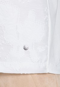 Blusa bianca ricamata con un motivo floreale. Orlo arrotondato con un dettaglio a bottone argentato di lato, realizzata in tessuto leggero.