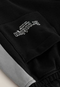 Pantalon en tissu noir et gris avec une poche patch noire affichant un texte blanc qui dit « "BT DUBLIN FUTURE SYSTEMS TWENTY FOUR SEVEN." »
