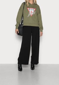 Olivově zelená zkrácená mikina s kapucí a logem, zkombinovaná s černými širokými kalhotami a černými kotníkovými botami. V ruce drží černou kabelku s detaily z síťoviny.