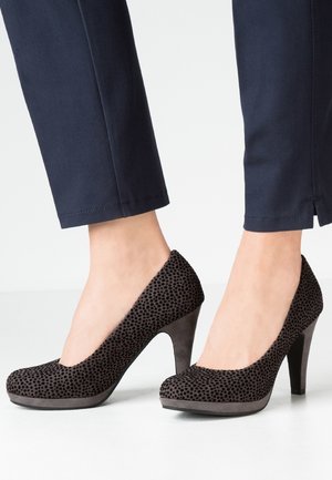 Chaussures noires à talons hauts avec un motif texturé, bout rond et un matériau lisse semblable à du daim sur un talon effilé, posées sur un pantalon marine.