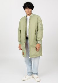 Man met krullend haar in een lange lichtgroene bomberjack, een wit shirt, lichtblauwe jeans en witte sneakers, staande tegen een witte achtergrond.