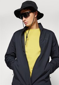 Schwarze leichte Jacke mit Reißverschluss, getragen über einem gelben strukturierten Hemd. Ein schwarzer Bucket Hat und dunkle Sonnenbrille vervollständigen den Look.