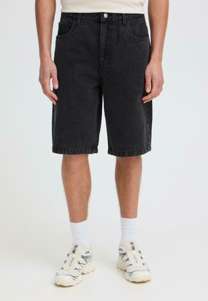Mann trägt schwarze Jeansshorts, weiße Socken und weiße Sportschuhe, steht vor einem schlichten hellen Hintergrund.