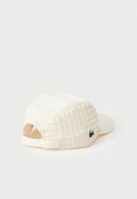 Krämfärgad keps i mjukt, texturerat tyg, med ett rutmönster på sidan och en grön logotypdetalj nära baksidan.
