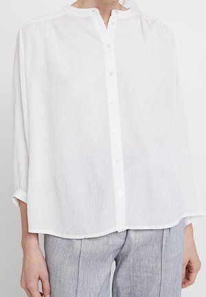 Kobieta w luźnej, białej koszuli na guziki z długim rękawem, w połączeniu z jasnoszarymi spodniami, stojąca na jednolitym tle.