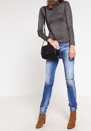 Femme portant un col roulé gris, un jean skinny bleu déchiré, des bottines en daim marron et un sac bandoulière noir avec un détail à pampille.