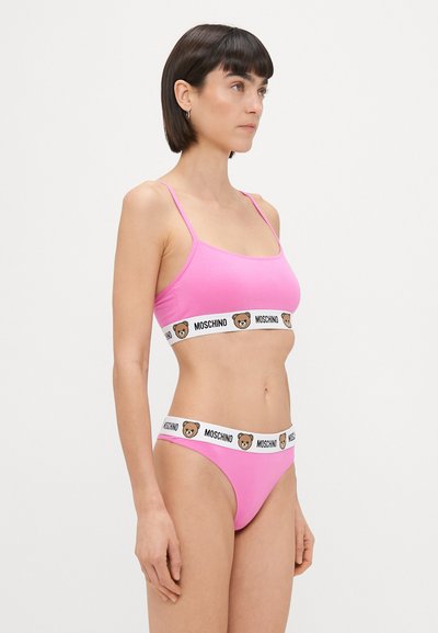 Rozā kokvilnas bralette un stringu komplekts ar plāniem siksniņām un baltu jostasvīru, uz kura drukāts Moschino lāča logo. Gluda tekstūra, vienkāršs dizains.