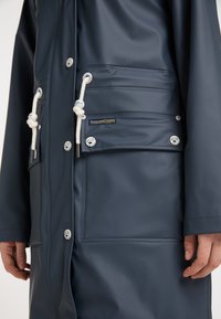 Marinblå regnjacka tillverkad av slätt, vattentätt material med tryckknappar, framficka och justerbara snoddar vid huvan och fållen.