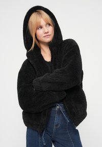 Μαύρο, αφράτο φλις μπουφάν με κουκούλα, μπροστινό φερμουάρ, φαρδιά μανίκια και άνετη εφαρμογή. Συνδυασμένο με ψηλόμεσο μπλε τζιν παντελόνι.