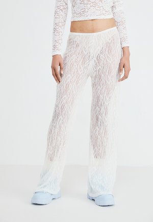Weiße Spitzenhose mit einem fließenden Design, das komplizierte wellenartige Muster und eine durchsichtige Textur aufweist; kombiniert mit hellblauen Sneakers.