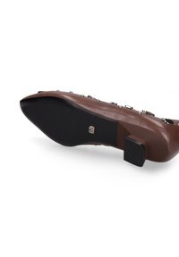 Chaussure en cuir marron avec un bout pointu, un petit talon carré, et des clous en argent décoratifs sur le côté. Semelle extérieure en caoutchouc noire.