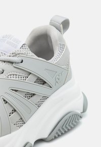 Baskets gris clair avec des panneaux en mesh, semelle en caoutchouc et accents en plastique. Branding notable sur le côté et finition texturée sur les détails.