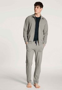 Lichtgrijze hoodie met rits en bijpassende joggers, beide in zachte stof. De joggers hebben een trekkoord en zakken. Zwarte t-shirt eronder.