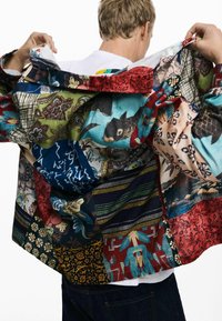 La chaqueta de patchwork multicolor presenta varios patrones, incluyendo diseños florales y abstractos, con una tela texturada y acentos de botones.
