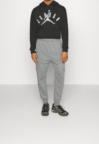 Svart huva med "JORDAN"-logotyp och grafik, grå avsmalnande mjukisbyxor med cargo-fickor, och svarta sportskor med gröna detaljer.