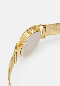 Montre en métal doré avec un boîtier rond et un bracelet en maille. Présente un cadran texturé et une couronne pour régler l'heure. Fini lisse à l'arrière.