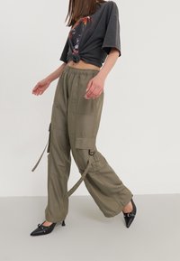 Olivgrüne Cargo-Hosen mit elastischem Bund, Seitentaschen und lockerer Passform. Getragen mit einem kurzen schwarzen T-Shirt mit Grafik und schwarzen High Heels.