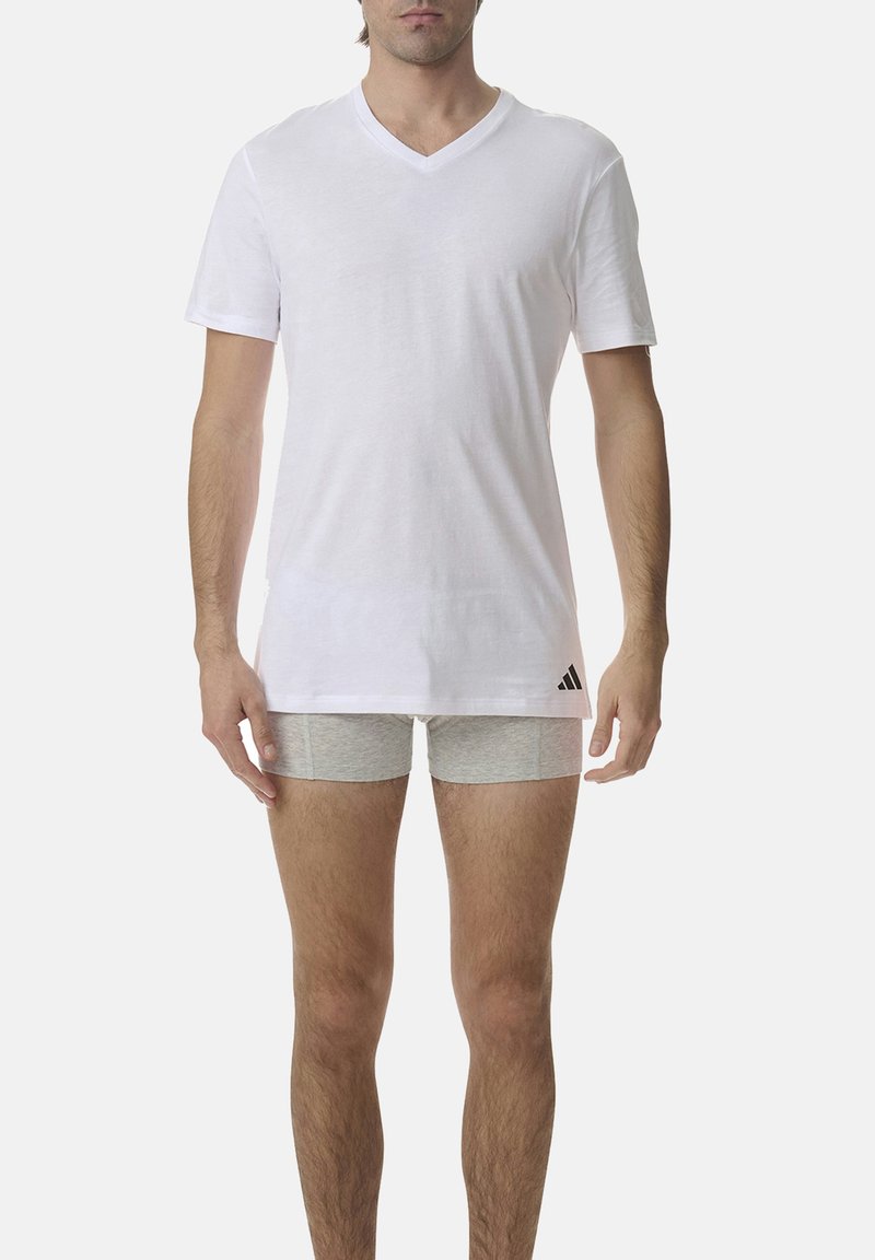 White cotton v-neck t-shirt with short sleeves, featuring a black Adidas logo on the lower left side. Model wears grey shorts.
