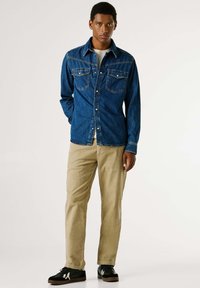 Traper košulja u plavoj boji s dva prednja džepa, kopčanje na gumbe i ovratnikom. Uparena s bež hlačama i crnim tenisicama.