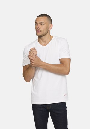 T-shirt en coton blanc à col en V et manches courtes, arborant un petit accent en forme de diamant rouge. Porté avec un jean foncé, affichant une coupe décontractée.