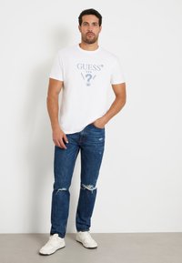T-shirt en coton blanc avec un imprimé graphique bleu, associé à un jean bleu déchiré et des baskets blanches, illustrant une tenue décontractée.