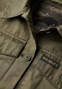 Olijfgroene blouse met een knoopsluiting, voorzien van twee borstzakken en een label. De binnenkant heeft een geborduurd ontwerp van een zwarte panter.