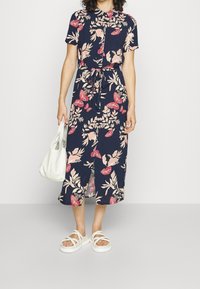 Abito midi blu navy floreale con maniche corte, colletto e vita con fiocco. Presenta motivi di farfalle e foglie. Abbinato a una borsa bianca e sandali.