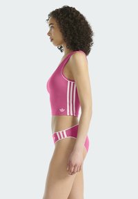 Różowy sportowy crop top o dopasowanym designie, z białymi akcentami w postaci trzech pasków po bokach oraz klasycznym logo na dolnym brzegu.