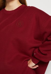 Sweat à col rond bordeaux, avec un logo brodé rond et des poignets côtelés. Fabriqué à partir d'un matériau doux et texturé.