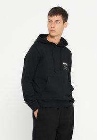 Haut à capuche en coton noir avec une poche kangourou, un capuchon à cordon et un logo imprimé sur la poitrine. Texture douce et coupe régulière.