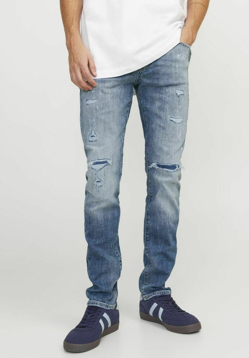 Vaqueros de mezclilla azul claro con un ajuste slim, detalles desgastados con desgarrones y un lavado desvanecido. Combinados con zapatillas oscuras.