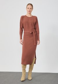 Une femme se tient debout vêtue d'une robe tricot couleur rouille à manches longues avec une ceinture à la taille et une fente sur le côté, associée à des bottes en daim beiges jusqu'aux genoux.