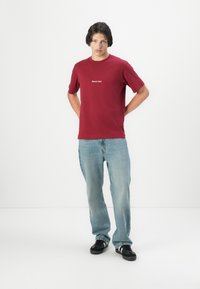 Rode katoenen T-shirt met korte mouwen, voorzien van een witte logo. Gecombineerd met lichtblauwe spijkerbroek en zwarte sneakers.
