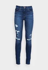 Jeans ajustados de mezclilla azul oscura con detalles desgastados en las rodillas y un diseño estándar de cinco bolsillos. Cuenta con un cierre de botón y pasadores para el cinturón.