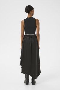 Robe sans manches noire avec un corsage ajusté et un ourlet asymétrique, présentant un tissu texturé et une ceinture en chaîne métallique à la taille.