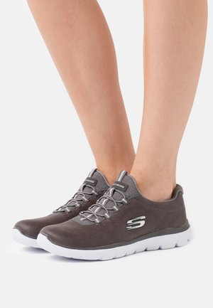 Zapatillas deportivas de ante gris con suelas blancas, con cordones grises y un logo plateado en el lateral. Punta redondeada y diseño bajo.