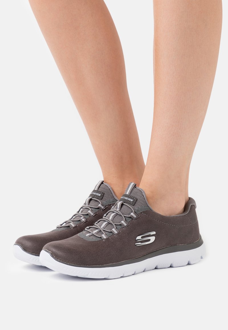 Baskets de sport en suède gris avec semelles blanches, dotées de lacets gris et d'un logo argenté sur le côté. Design à bout rond et coupe basse.