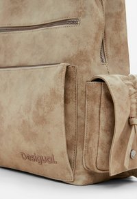 Sac à dos en simili cuir beige avec finition texturée, poche zippée sur le devant, poche latérale et logo "Desigual" en relief. Design minimaliste et fonctionnalités pratiques.