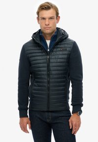 Heren hooded jacket, combinatie van gewatteerd donkerblauw en zacht textiel, met ritszakken en subtiele branding op de borst.