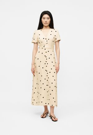 Mujer de pie con un vestido midi beige con lunares negros, botones delanteros, mangas cortas, un cinturón y sandalias negras de punta abierta.