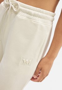 Pantalones deportivos de color crema hechos de una tela suave, con una cinturilla elástica, cordón, bolsillos laterales y un sutil logo embosado.