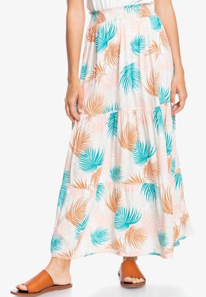 Jupe maxi fluide avec imprimé de feuilles de palmier bleu-vert et orange portée avec des sandales marron à bout ouvert sur fond blanc.