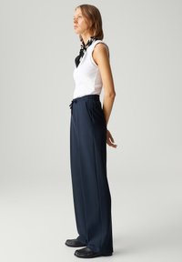 Navy wijde broek met elastische tailleband, zijplooien en een trekkoord, gedragen met een aansluitend wit mouwloos topje en een zwart-wit sjaal.