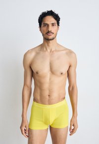 Gule bomuldsboxer shorts med en tætsiddende pasform og hvid elastisk talje, der viser mærkenavnet "BOSS." Modellen står med afslappede arme.