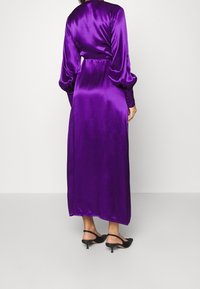 Lila Satin-Maxikleid mit langen, gepufften Ärmeln und Knopfdetails. Glatte Textur mit taillierter Passform und fließendem Schnitt.