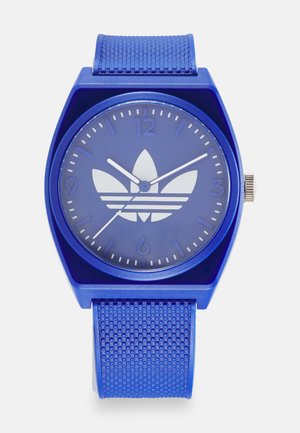 Montre numérique bleue avec un boîtier en plastique carré, un bracelet en silicone texturé, un logo blanc et des aiguilles des minutes/heures positionnées sur le cadran rond.