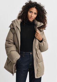 Puffy beige winterjas met hoge kraag en ritssluiting, voorzien van zijzakken en een gequilte textuur, gedragen over een zwarte coltrui.