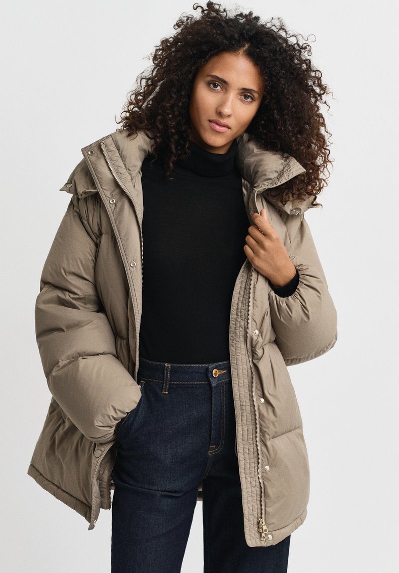 Puffy beige winterjas met hoge kraag en ritssluiting, voorzien van zijzakken en een gequilte textuur, gedragen over een zwarte coltrui.