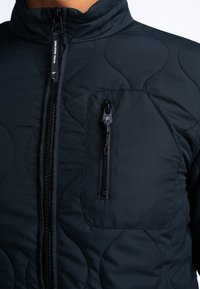 Schwarze gesteppte Jacke mit hohem Kragen, Frontreißverschluss und einer kleinen Brusttasche mit Reißverschluss. Strukturiertes Muster über die gesamte Jacke.